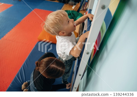 Climbing the ladder that is on the wall. Preschool child with teacher during developmental activities in a playroom Climbing the ladder that is on the wall. Preschool child with teacher during developmental activities in a playroom 139244336