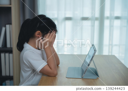 Young girl feeling frustration and sadness while looking at a tablet device on a wooden desk in a well-lit room with soft curtains in the background Young girl feeling frustration and sadness while looking at a tablet device on a wooden desk in a well-lit room with soft curtains in the background 139244423