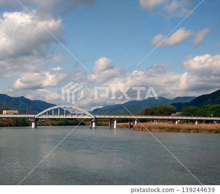 An arch bridge spanning a river flowing beneath a blue sky and clouds, and a view of the surrounding mountains. An arch bridge spanning a river flowing beneath a blue sky and clouds, and a view of the surrounding mountains. 139244639