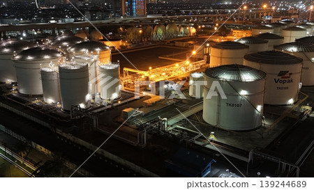 High-angle night view of a fuel storage tank terminal with multiple large, illuminated cylinders and a complex piping infrastructure at an industrial port facility, glowing in the dark. High-angle night view of a fuel storage tank terminal with multiple large, illuminated cylinders and a complex piping infrastructure at an industrial port facility, glowing in the dark. 139244689