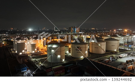 High-angle night view of a fuel storage tank terminal with multiple large, illuminated cylinders and a complex piping infrastructure at an industrial port facility, glowing in the dark. High-angle night view of a fuel storage tank terminal with multiple large, illuminated cylinders and a complex piping infrastructure at an industrial port facility, glowing in the dark. 139244692