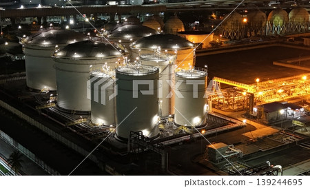High-angle night view of a fuel storage tank terminal with multiple large, illuminated cylinders and a complex piping infrastructure at an industrial port facility, glowing in the dark. High-angle night view of a fuel storage tank terminal with multiple large, illuminated cylinders and a complex piping infrastructure at an industrial port facility, glowing in the dark. 139244695