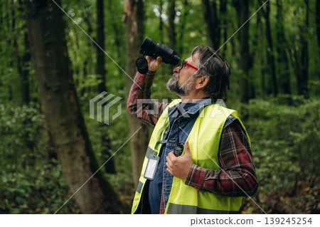 Far away, looking through the binoculars. Senior forest ecologist is working in the woods. In the uniform Far away, looking through the binoculars. Senior forest ecologist is working in the woods. In the uniform 139245254