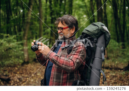 Camera is in hands, making photos. Hiking experience, senior man is in the forest at daytime Camera is in hands, making photos. Hiking experience, senior man is in the forest at daytime 139245276
