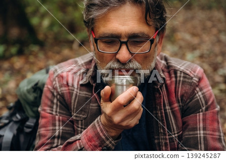 Warm drink in hands, thermos. Hiking experience, senior man is in the forest at daytime Warm drink in hands, thermos. Hiking experience, senior man is in the forest at daytime 139245287