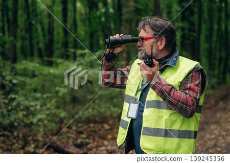 Radio walkie talkie and binoculars in hands. Senior forest ecologist is working in the woods. In the uniform 139245356