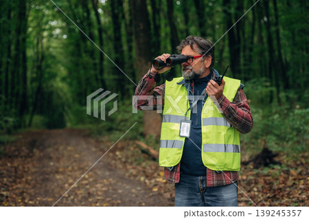 Radio walkie talkie and binoculars in hands. Senior forest ecologist is working in the woods. In the uniform Radio walkie talkie and binoculars in hands. Senior forest ecologist is working in the woods. In the uniform 139245357