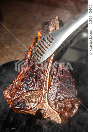 Grilled t-bone steak being flipped with tongs on hot pan, juicy seared crust 139245462