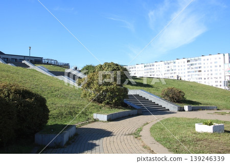 Urban garden with winding paths. Sunlit park featuring curved trails and lush greenery Urban garden with winding paths. Sunlit park featuring curved trails and lush greenery 139246339