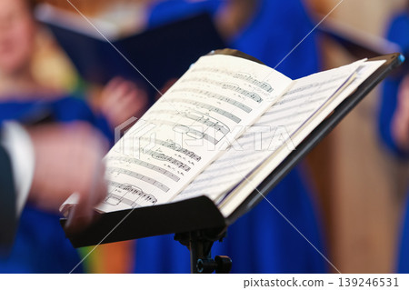 Open sheet music on a stand as a conductor leads a choir Open sheet music on a stand as a conductor leads a choir 139246531