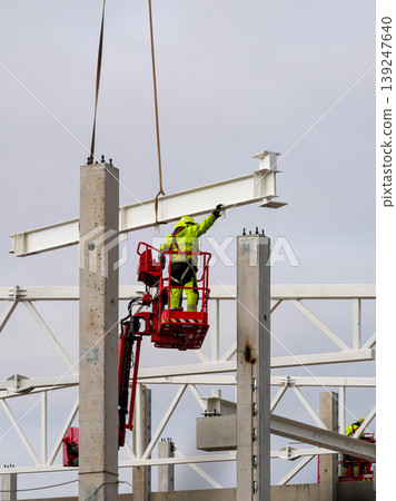 Construction worker guiding suspended steel beam into place on concrete columns with crane Construction worker guiding suspended steel beam into place on concrete columns with crane 139247640