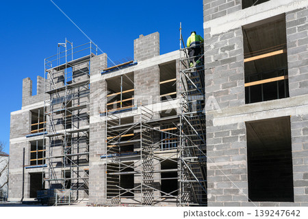 Worker on scaffolding at concrete block building under construction with open floors and blue sky Worker on scaffolding at concrete block building under construction with open floors and blue sky 139247642