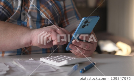 A person types on a smartphone while holding a calculator in one hand. Papers and a pen are on the table. The time appears to be during the day indoors. A person types on a smartphone while holding a calculator in one hand. Papers and a pen are on the table. The time appears to be during the day indoors. 139247975