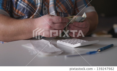 A person counts banknotes while seated at a table with a calculator nearby in a dimly lit room during the late afternoon. Notes and a pen are also present on the table. 139247982