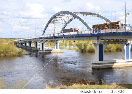 Arched metal bridge over a river with heavy trucks transporting timber and cargo. Modern road infrastructure, logistics, and transportation concept. Arched metal bridge over a river with heavy trucks transporting timber and cargo. Modern road infrastructure, logistics, and transportation concept. 139248051