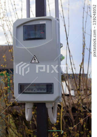 Outdoor electric meter box mounted on a pole in a rural area. Grey plastic electrical panel with high voltage warning sign, circuit breakers 139248052