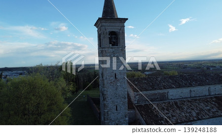 A scenic view of the medieval Pieve di San Pancrazio in Montichiari, Italy. The stone church with its bell tower is surrounded by a green park with cypress trees under a clear sky. 139248188