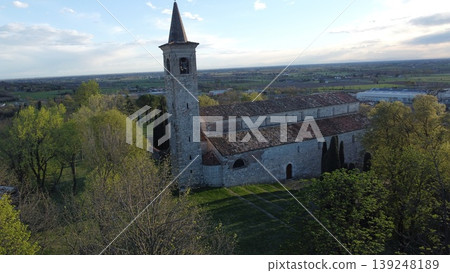 A scenic view of the medieval Pieve di San Pancrazio in Montichiari, Italy. The stone church with its bell tower is surrounded by a green park with cypress trees under a clear sky. 139248189