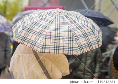 Close up of beige plaid umbrella with rain drops. Plaid umbrella shields woman in crowded rainy city street. Concept of umbrella protection, urban travel, wet weather Close up of beige plaid umbrella with rain drops. Plaid umbrella shields woman in crowded rainy city street. Concept of umbrella protection, urban travel, wet weather 139248284