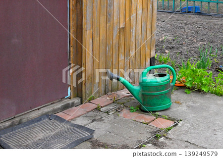 green plastic watering can for plants stands on the ground near the wooden wall of a shed green plastic watering can for plants stands on the ground near the wooden wall of a shed 139249579