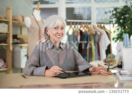 Positive mature female dressmaker drawing in tablet while sitting at table Positive mature female dressmaker drawing in tablet while sitting at table 139251773