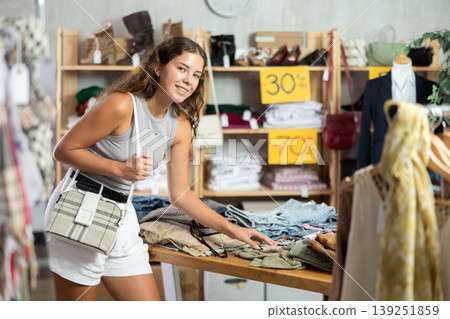Positive young woman choosing trousers in clothing store Positive young woman choosing trousers in clothing store 139251859