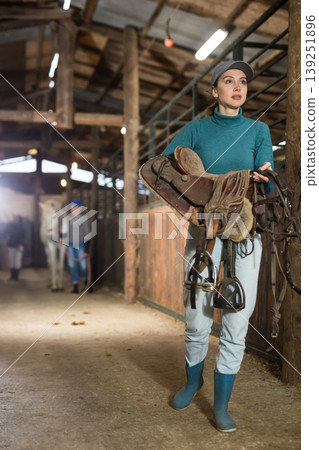 Female rancher carrying horse tack and saddle in horse farm Female rancher carrying horse tack and saddle in horse farm 139251896