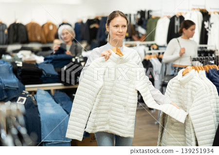 Positive woman chooses white winter down jacket in clothing store Positive woman chooses white winter down jacket in clothing store 139251934