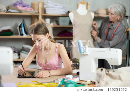 Female tailor drawing design on tablet with mature female worker in background Female tailor drawing design on tablet with mature female worker in background 139251952