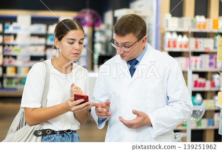 Young girl showing to male pharmacist something displayed on her smartphone Young girl showing to male pharmacist something displayed on her smartphone 139251962