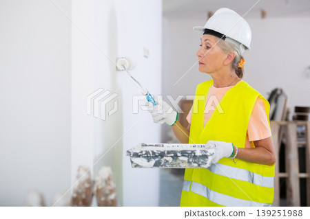 Portrait of elderly positive woman painter in protective helmet and yellow vest who paints walls with paint roller 139251988
