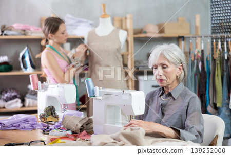 Mature tailoress using sewing-machine while other working with mannequin Mature tailoress using sewing-machine while other working with mannequin 139252000