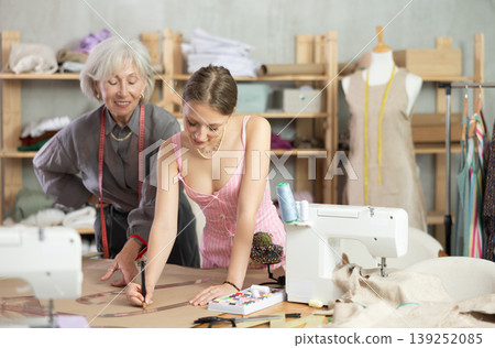 Elderly woman seamstress teaches young woman to draw pattern on paper 139252085