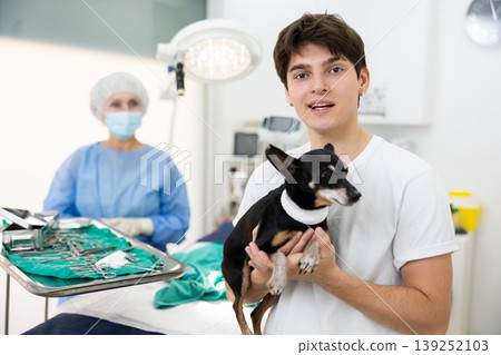 Young man owner hold and cuddles Miniature Pinscher pet after manipulations in veterinary clinic Young man owner hold and cuddles Miniature Pinscher pet after manipulations in veterinary clinic 139252103