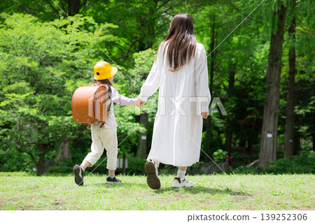 A mother and her daughter, wearing a schoolbag, walk hand in hand through a park surrounded by fresh greenery. A full-body shot of a mother and daughter going to school, with their faces obscured from behind. A mother and her daughter, wearing a schoolbag, walk hand in hand through a park surrounded by fresh greenery. A full-body shot of a mother and daughter going to school, with their faces obscured from behind. 139252306