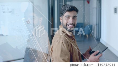 Leaning Indian man holding tablet and glancing in office corridor with glass partition, copy space Leaning Indian man holding tablet and glancing in office corridor with glass partition, copy space 139252944
