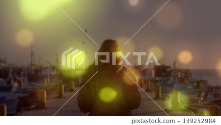 Adult female watching horizon wearing padded coat on harbor pier at sunset, with moored boats Adult female watching horizon wearing padded coat on harbor pier at sunset, with moored boats 139252984