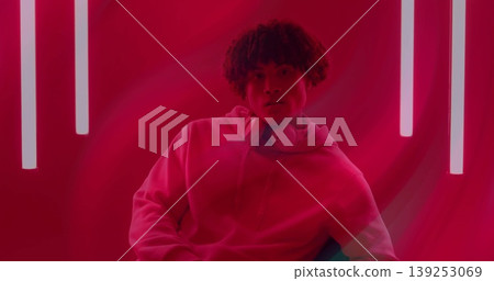 Sitting man wearing hoodie and over-ear headphones on chair in red studio, with vertical neon tubes Sitting man wearing hoodie and over-ear headphones on chair in red studio, with vertical neon tubes 139253069