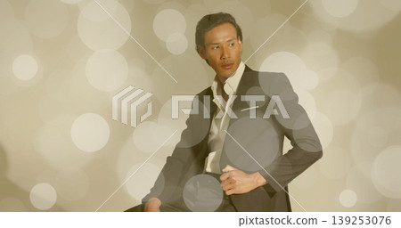 Posing adult Asian man on stool adjusting suit jacket and dress shirt in studio, bokeh backdrop Posing adult Asian man on stool adjusting suit jacket and dress shirt in studio, bokeh backdrop 139253076