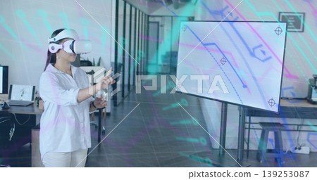 Pointing Asian woman wearing VR headset selecting virtual elements in open-plan office, copy space 139253087