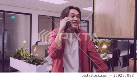 Leaning Asian man in white T-shirt, red shirt speaking on smartphone in office by string-lit plant Leaning Asian man in white T-shirt, red shirt speaking on smartphone in office by string-lit plant 139253088