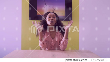 Host wearing pink blouse gesturing toward camera at table in studio set, with wind turbine artwork 139253090