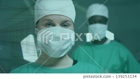 Standing woman surgeon wearing green gown and surgical mask in operating room, with overhead lights Standing woman surgeon wearing green gown and surgical mask in operating room, with overhead lights 139253134