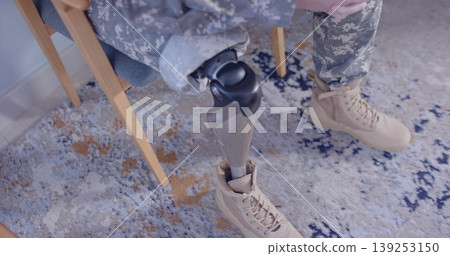 Sitting soldier wearing camo pants, tan boot on living-room rug, with wooden chair, prosthetic limb 139253150