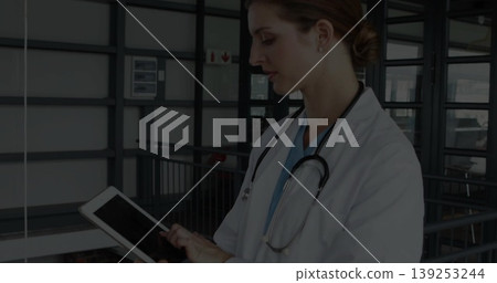 Reviewing doctor in blue scrubs in white coat using tablet on hospital corridor with stethoscope Reviewing doctor in blue scrubs in white coat using tablet on hospital corridor with stethoscope 139253244