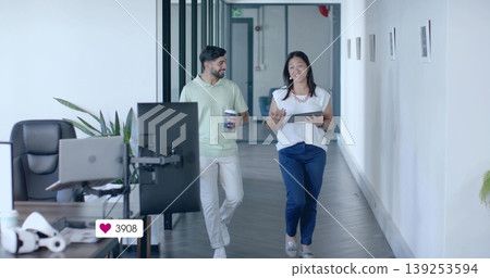 Walking colleagues conversing in hallway, with tablet, coffee cup, monitors, VR headset and plants Walking colleagues conversing in hallway, with tablet, coffee cup, monitors, VR headset and plants 139253594