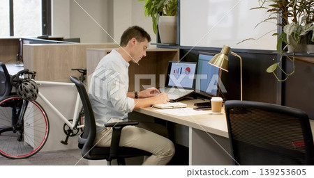 Writing man in shirt reviewing laptop charts at office desk with coffee cup and bicycle Writing man in shirt reviewing laptop charts at office desk with coffee cup and bicycle 139253605