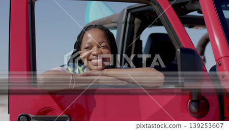 Leaning woman wearing tie-dye T-shirt resting on red SUV on beach, with surfboard on roof rack Leaning woman wearing tie-dye T-shirt resting on red SUV on beach, with surfboard on roof rack 139253607