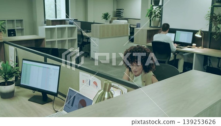 Curly-haired worker in sleeveless top reviewing charts on monitor at desk with lamp, copy space 139253626