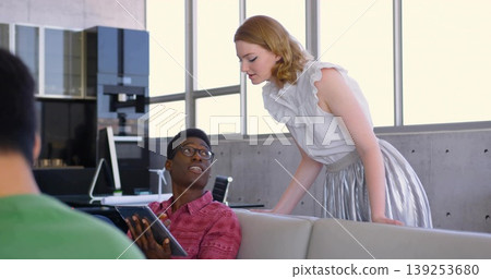 Leaning woman in business attire speaking with seated man holding tablet on sofa in office lounge 139253680